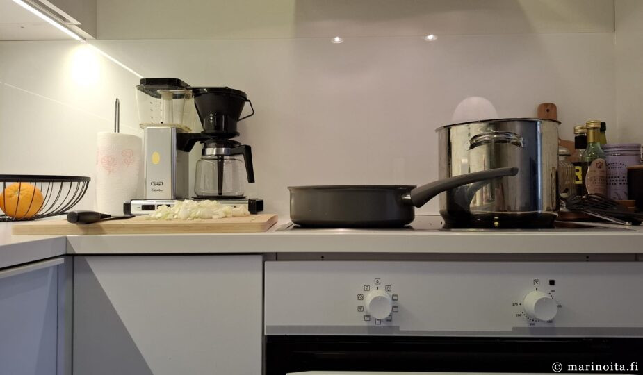 Liedellä keraaminen paistokasari ja teräskattila ja vieressä puinen leikkuulauta.